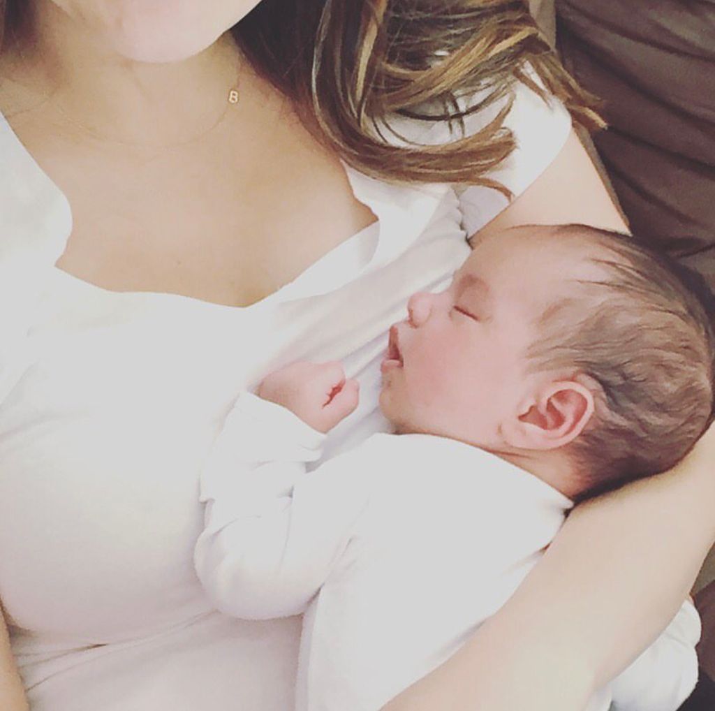 A woman holding a baby and wearing a gold necklace with a B letter charm.