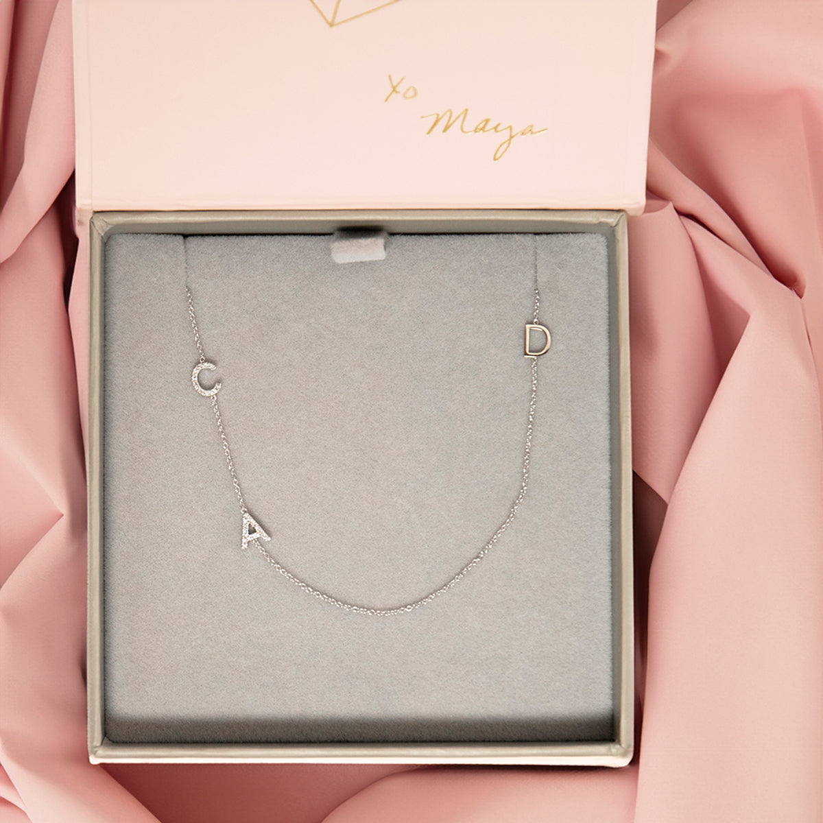 A Custom Gold Necklace in white gold with three letters, a pavé C and A on the left side and a D on the right side, in a Maya Brenner case against a pink fabric background.