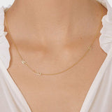 A close-up of a woman’s neck wearing a Custom Gold Necklace in yellow gold with three letter charms, a K and an E on the left side and a B on the right side, and a white blouse.