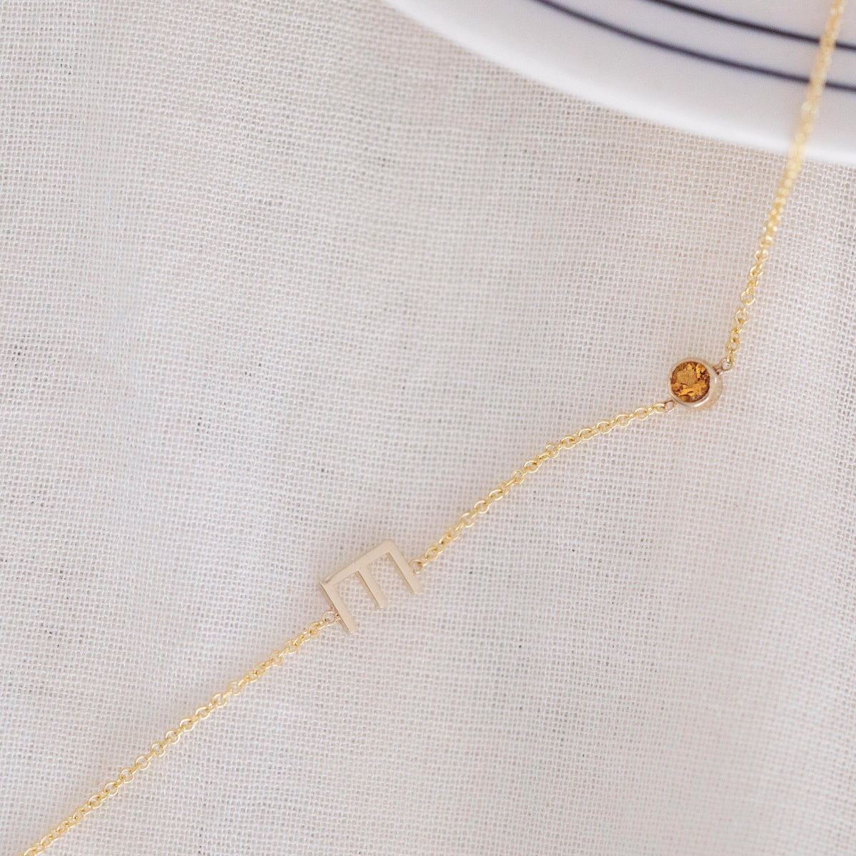 A Custom Gold Necklace in yellow gold with a letter E charm and a birthstone, on an off-white fabric background.