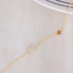 A Custom Gold Necklace in yellow gold with a letter E charm and a birthstone, on an off-white fabric background.