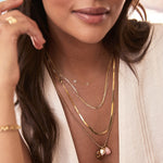 Close-up of a woman wearing multiple yellow gold necklaces, including a Custom Gold Necklace with the letters K and R, an Element Short Link Chain, an Overboard Layering Chain, and a Round Curb Chain with a Pink Opal Tear Drop Charm, an Engraved Heart Charm, and a Diamond Heart Locket Charm, paired with a cream top.