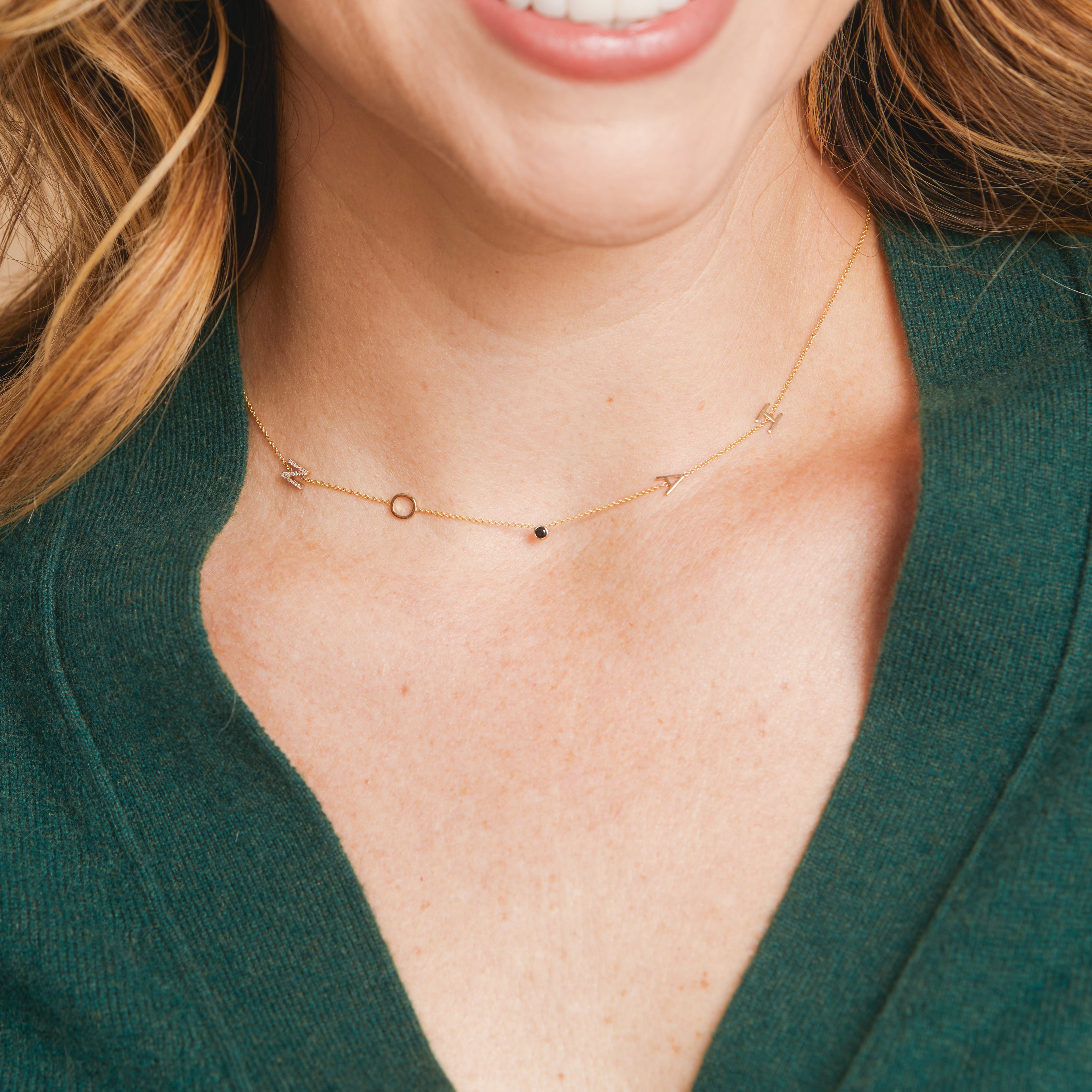 Close-up of a person wearing a delicate gold necklace with small lettrers and a black stones.