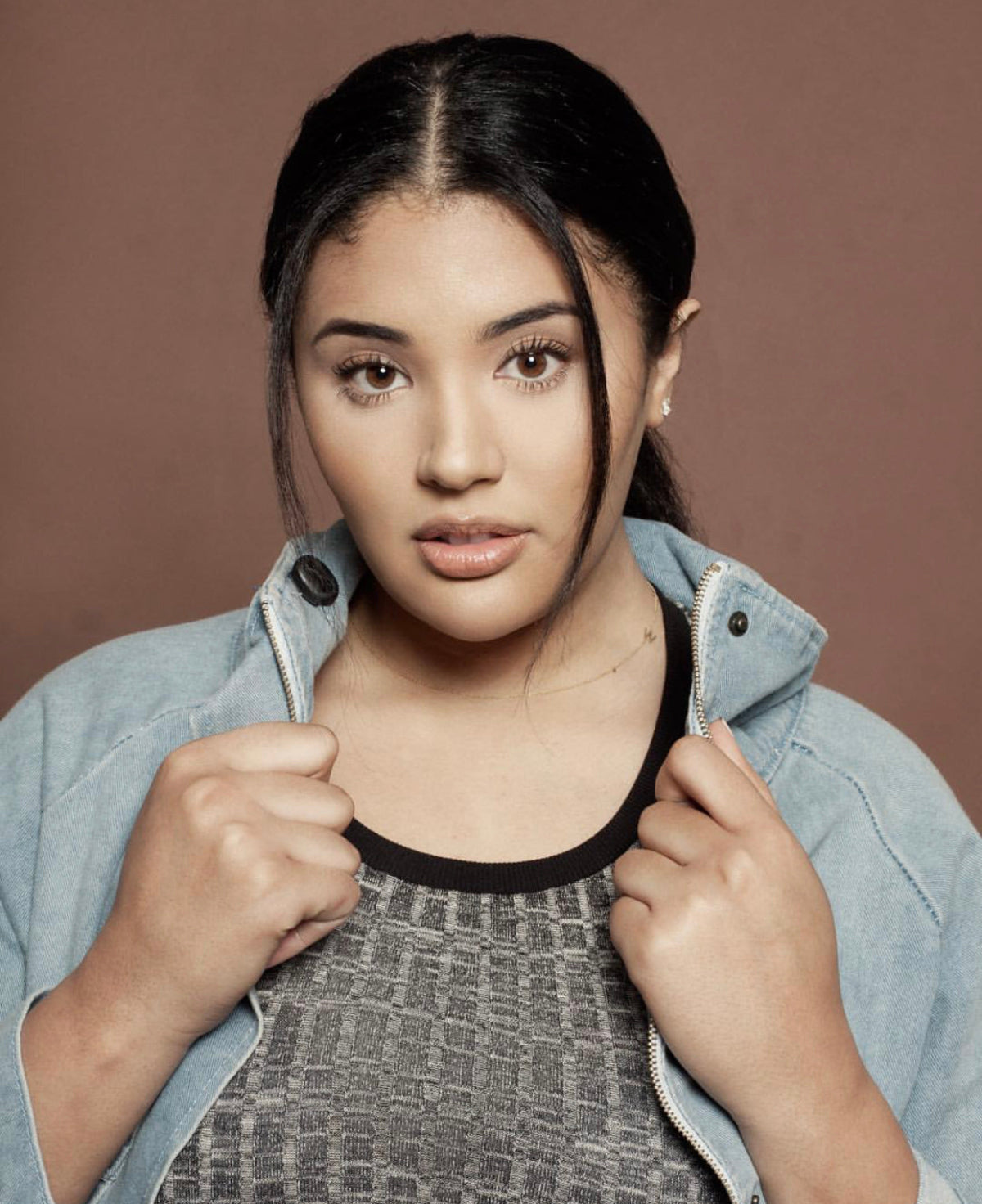 A woman wearing a dark grey top and denim jacket, a necklace with a letter W charm and stud earrings.