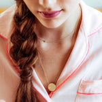 A close-up of a woman wearing a pink shirt and two gold necklaces - a short one with a letter A charm and a long one with a gold solid circle.