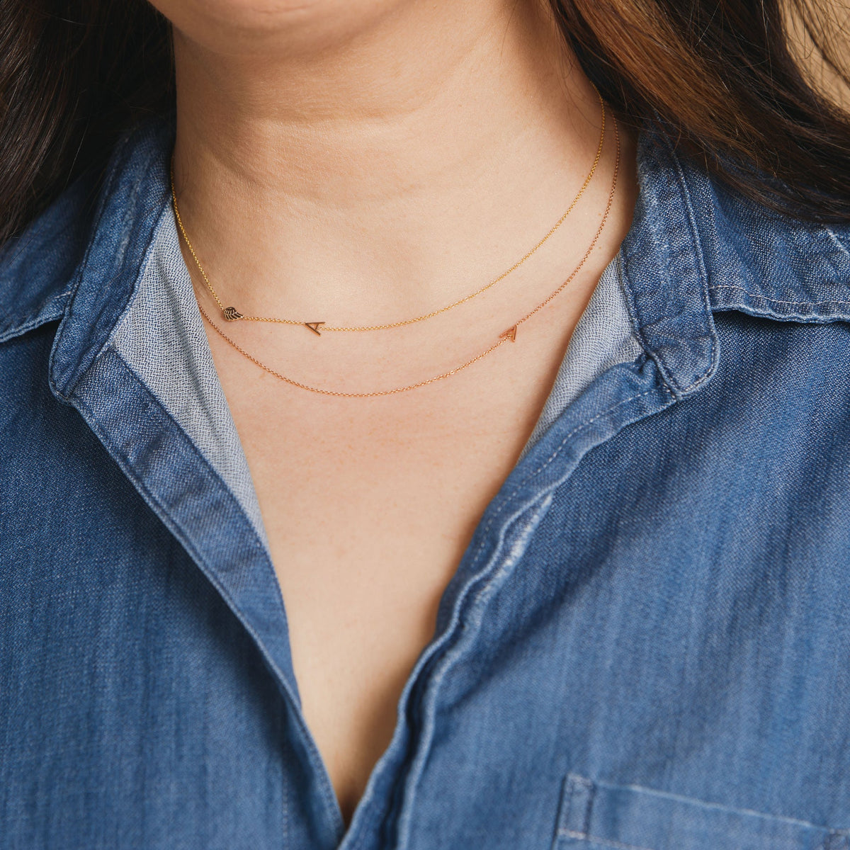 Close-up of a woman wearing two necklaces - one in rose gold with a letter A charm, and one in gold with a letter A charm and an angel wing.