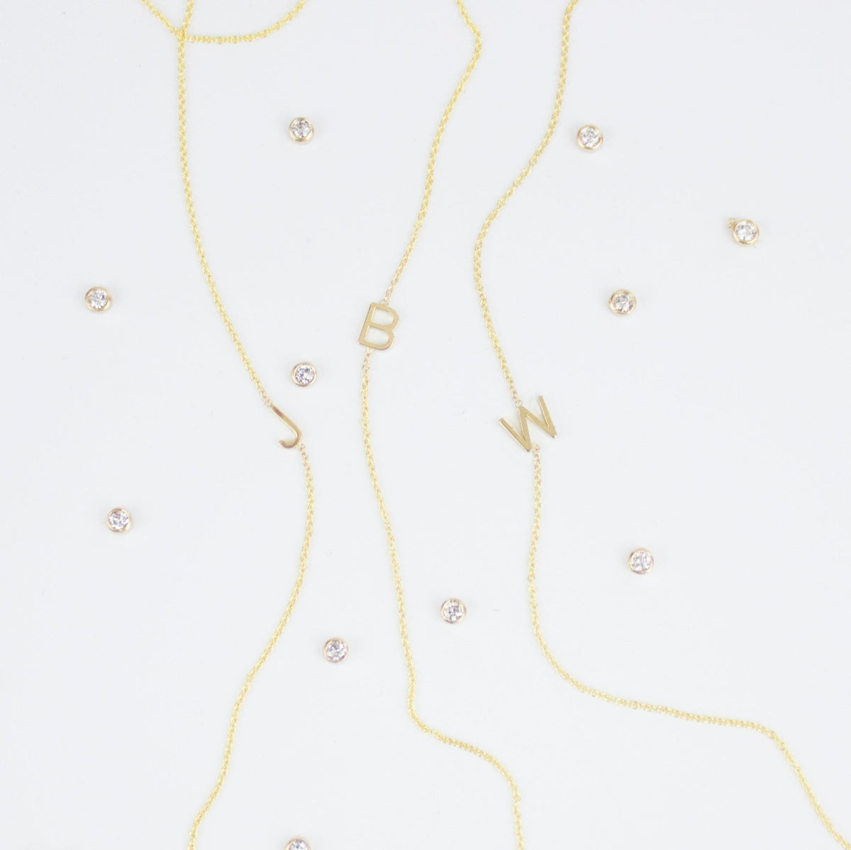 Three gold necklaces with single letter charms, being J, B and W, featuring multiple little gemstones on a white background.