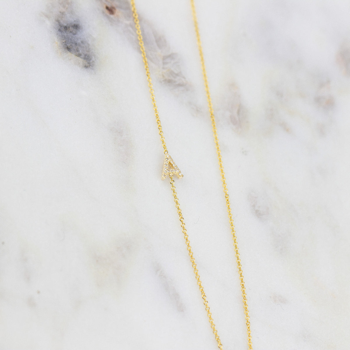 A gold necklace with a pave letter A charm  on a marble background.