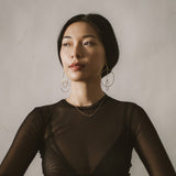 A woman in a black sheer long sleeve top wearing a yellow gold necklace with a letter S charm and drop earrings with circles.