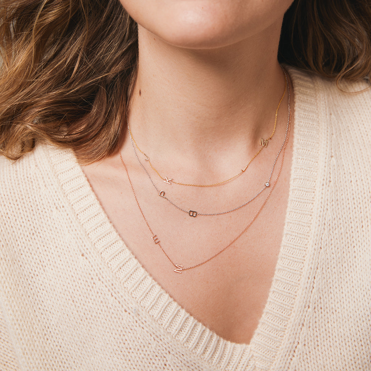 A close-up of a woman’s neck wearing three custom gold necklaces: a four-letter yellow gold necklace with the letters J, K, L and M, a white gold necklace with the letters P and B and a birthstone, and a two-letter rose gold necklace with the letters E and M, and a cream knit.