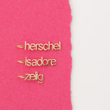 Three yellow gold Petite Nameplate Pendants with 'herschel', 'isadore', and 'zelig' on a pink and white background. 