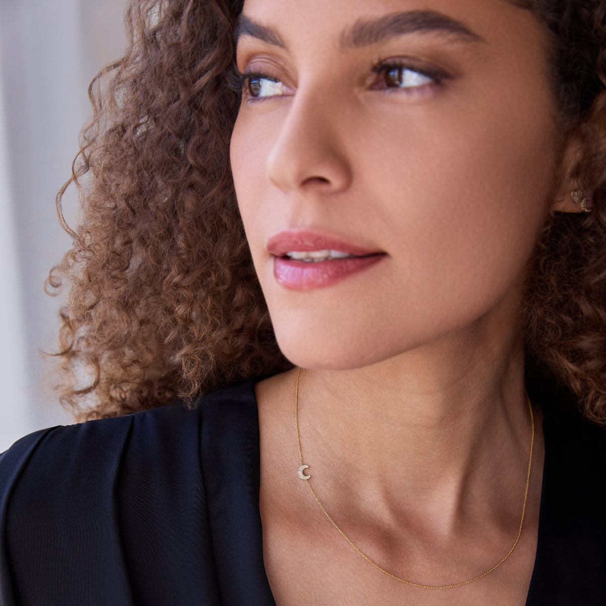 A woman wearing a yellow gold 14k Gold Asymmetrical Charm Necklace - Pavé Moon, with curly hair, styled in a navy top.