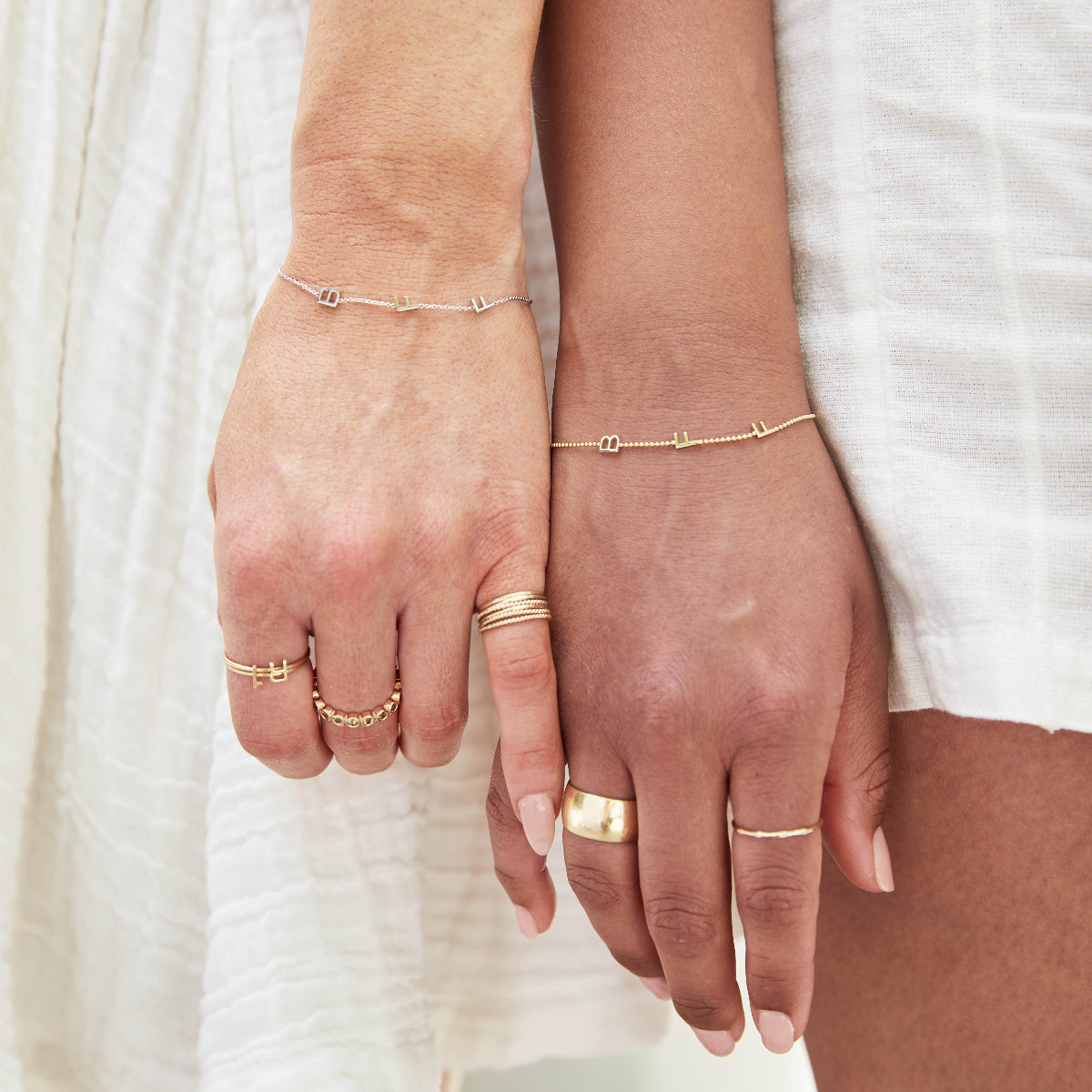 Close-up of two hands, each wearing a BFF Bracelet and several yellow gold rings, including a 14K Gold Letter Ring in A, a Lucky Number Ring in 1, a Hydrangea Bouquet Ring, a Thick Band, Delicate Bands, Delicate Twist Bands and a 4 Diamond Ring, against a white fabric background. 