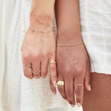 Close-up of two hands, each wearing a BFF Bracelet and several yellow gold rings, including a 14K Gold Letter Ring in A, a Lucky Number Ring in 1, a Hydrangea Bouquet Ring, a Thick Band, Delicate Bands, Delicate Twist Bands and a 4 Diamond Ring, against a white fabric background. 