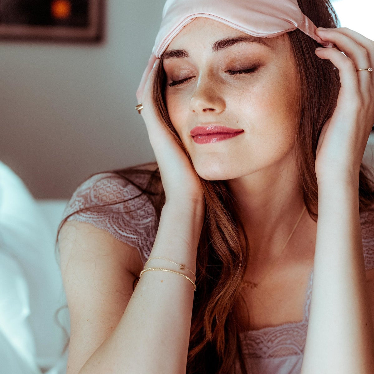 Gold bracelet with an M letter charm shown on a woman wearing a pink satin headband and lace lingerie, sitting on a bed.