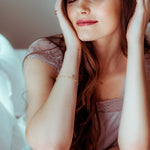Gold bracelet with an M letter charm shown on a woman wearing a pink satin headband and lace lingerie, sitting on a bed.