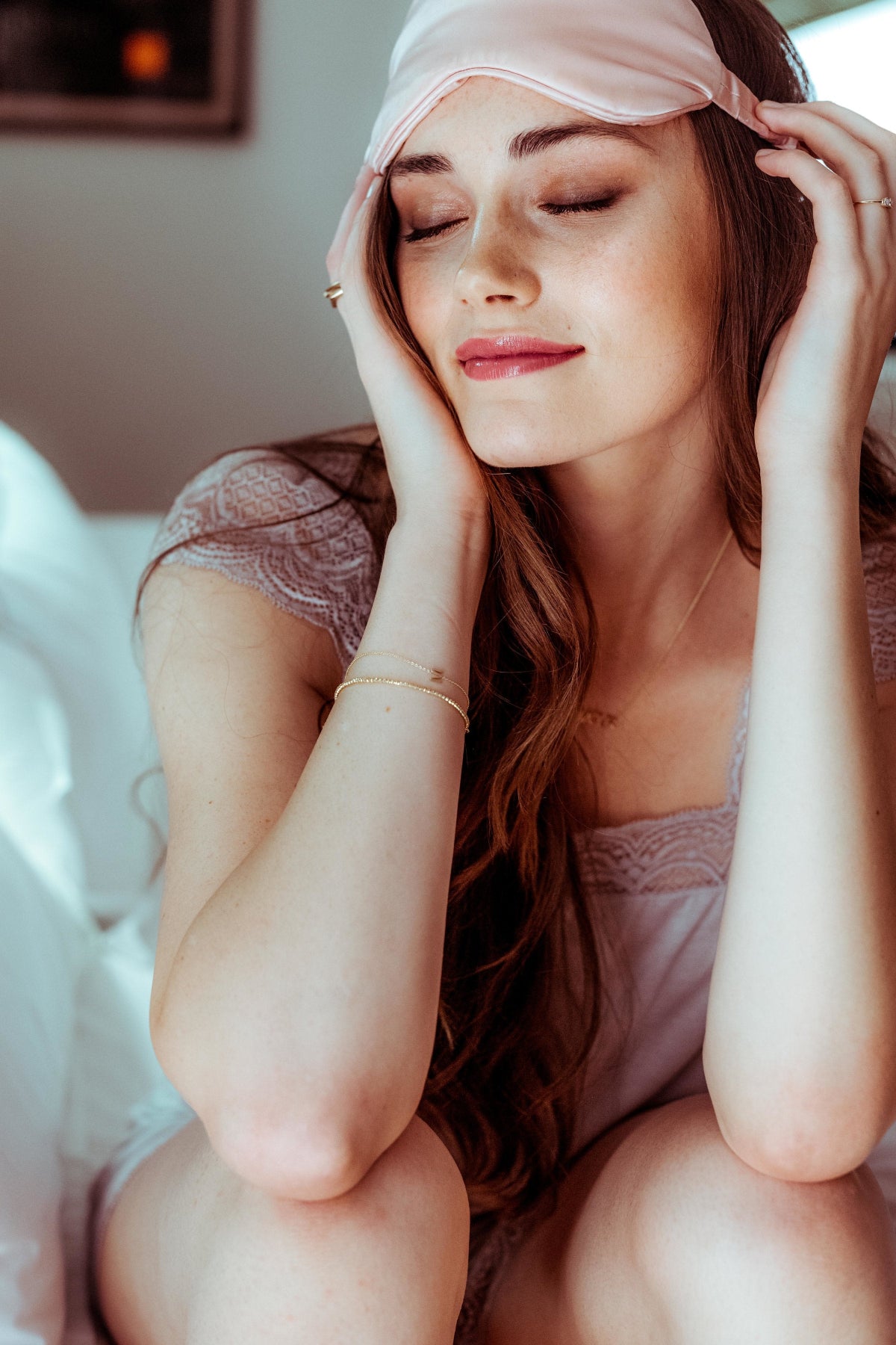 Gold bracelet with an M letter charm shown on a woman wearing a pink satin headband and lace lingerie, sitting on a bed.