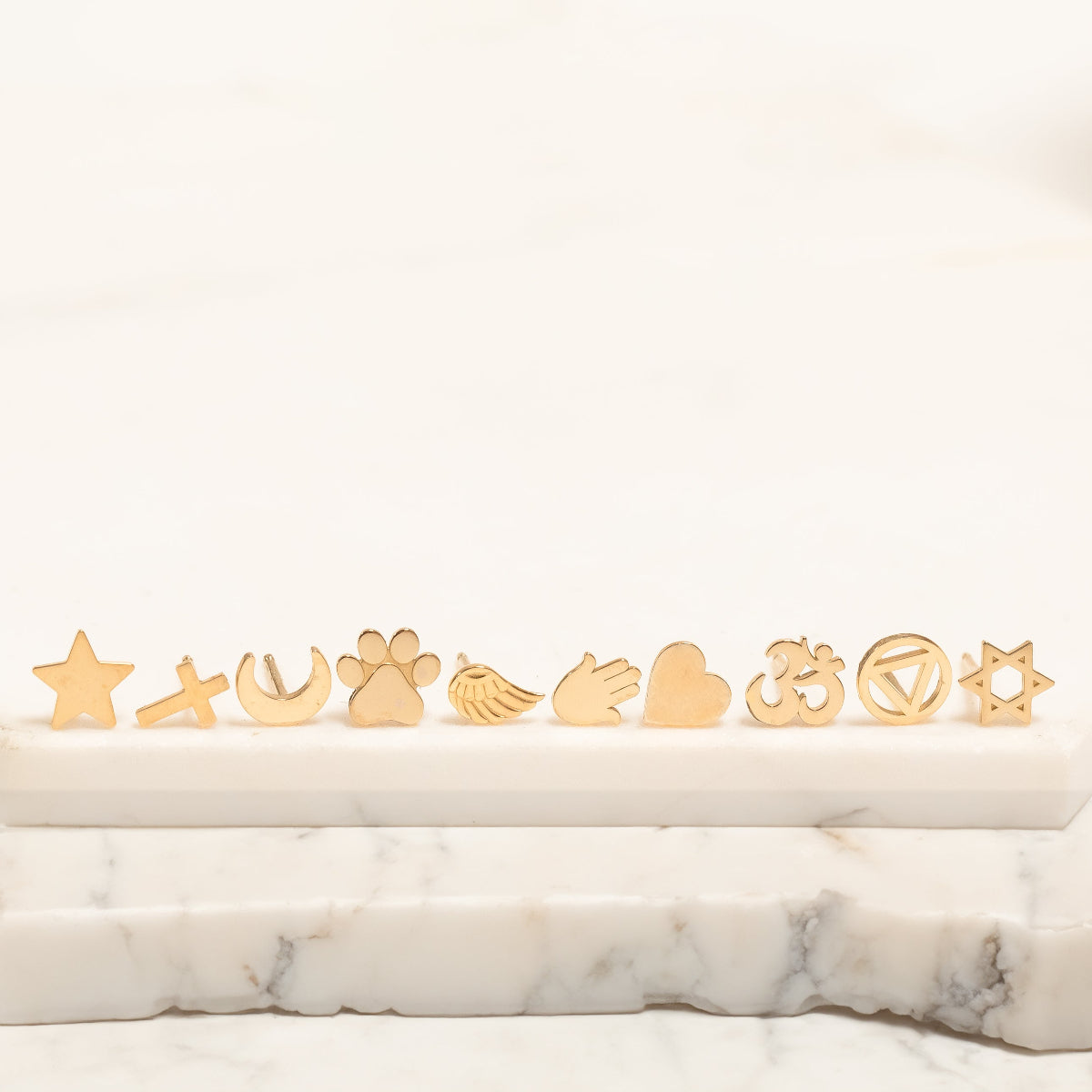 Several Charm Studs – Star, Cross, Moon, Paw, Angel Wing, Hamsa, Heart, Om, Recovery and Star of David laid on a marble stone surface and a white background. 