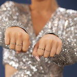 A pair of hands in fists wearing two Pave Diamond Rings, two Open Diamond Ring and a 4 Diamond Ring, and a silver sequenced top with a blurred background. 