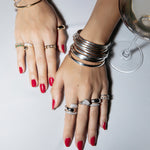 A pair of hands wearing several white gold For Keeps Bangle and Bouquet Rings two Make it a Double Rings, two Cosmo Rings, a C'est Moi Link Ring, two Martini Rings, a Braided Ring, a Love on the Rocks Ring, a Flirt Bangle, with red nails next to a cocktail glass against a light grey background. 