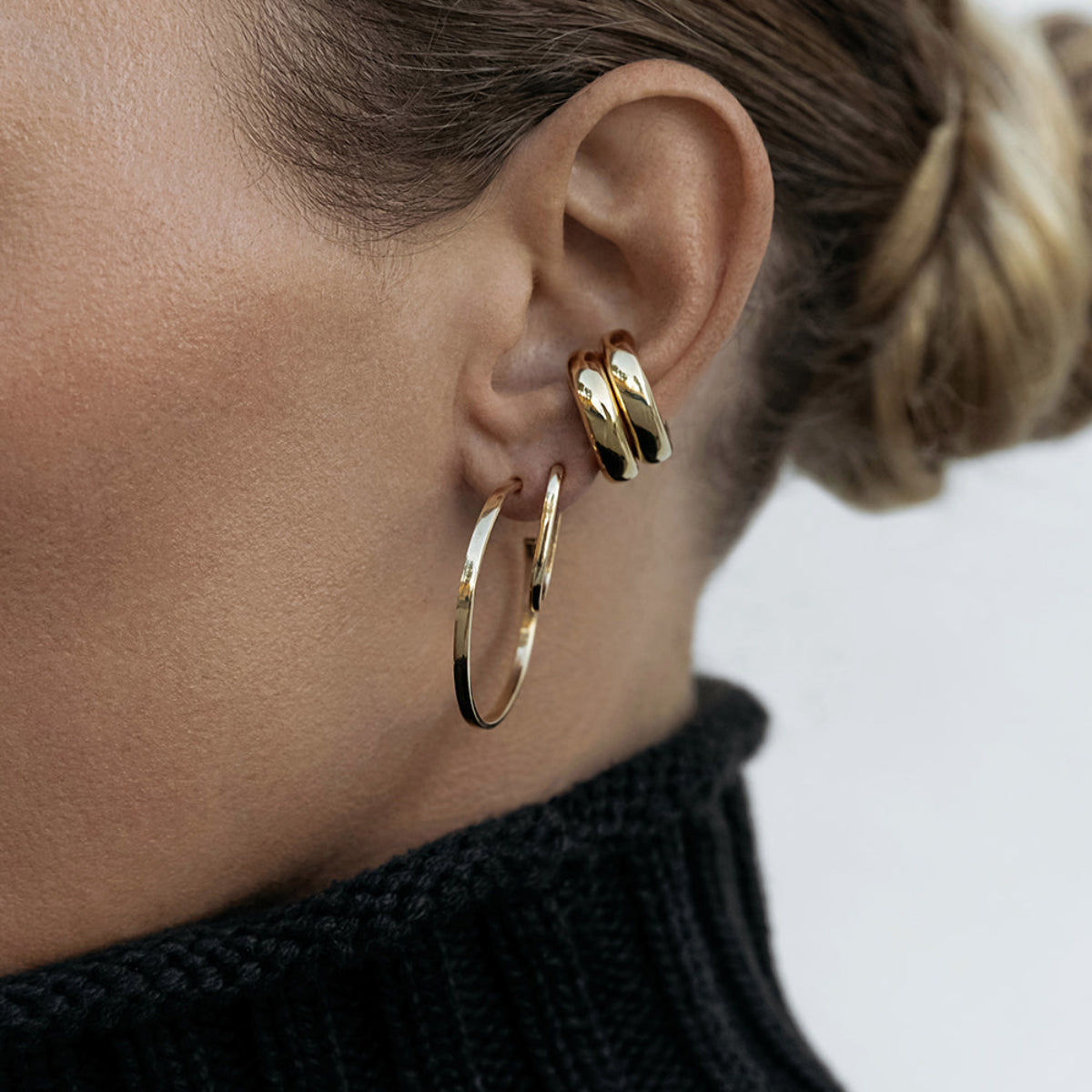 Close-up of an ear wearing two Ready to Mingle Ear Cuffs, an Oval Hoop and a Sleek & Chic Hoop - Medium in yellow gold with a blurred background.  