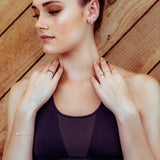 A woman wearing a yellow gold Custom Gold Bracelet with a heart charm, several gold rings, a necklace and earrings, in a dark purple sports bra against a wooden background.