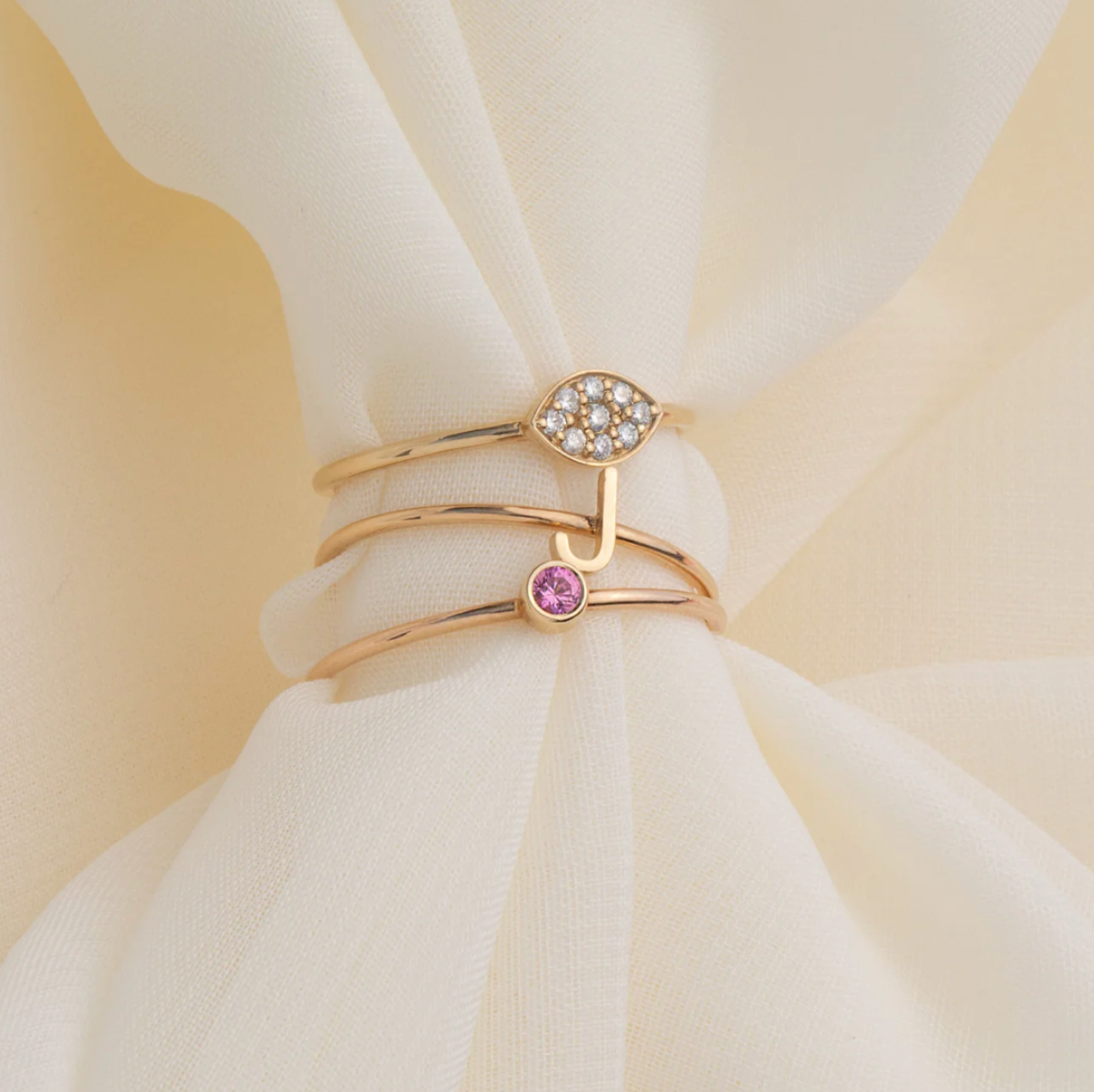 Three gold rings, including a yellow gold Pavé Petal Ring, a rose gold J Letter Ring, and a rose gold Birthstone Ring on a cream fabric background. 