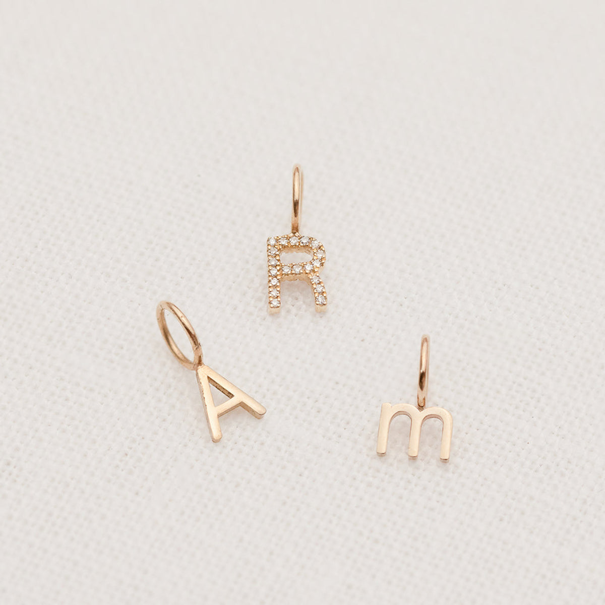 A Petite Letter Charm in m, Gold Letter Charm in A and a Pave Letter Charm R on a white textured background. 