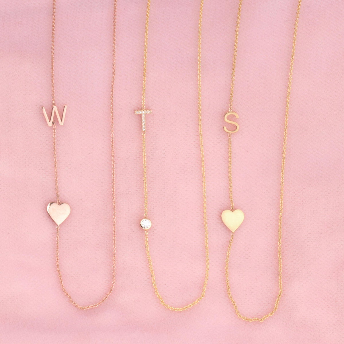 Three necklaces against a pink background -  Monogram Necklace with Heart Rose Gold and a W letter charm, a  Pavé Monogram Necklace with Diamond and a T letter charm, and a Monogram Necklace with Heart Yellow Gold and an S letter charm. 
