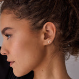 A close-up of a woman's side profile wearing two earrings - a mini letter E gold stud and a diamond stud. 