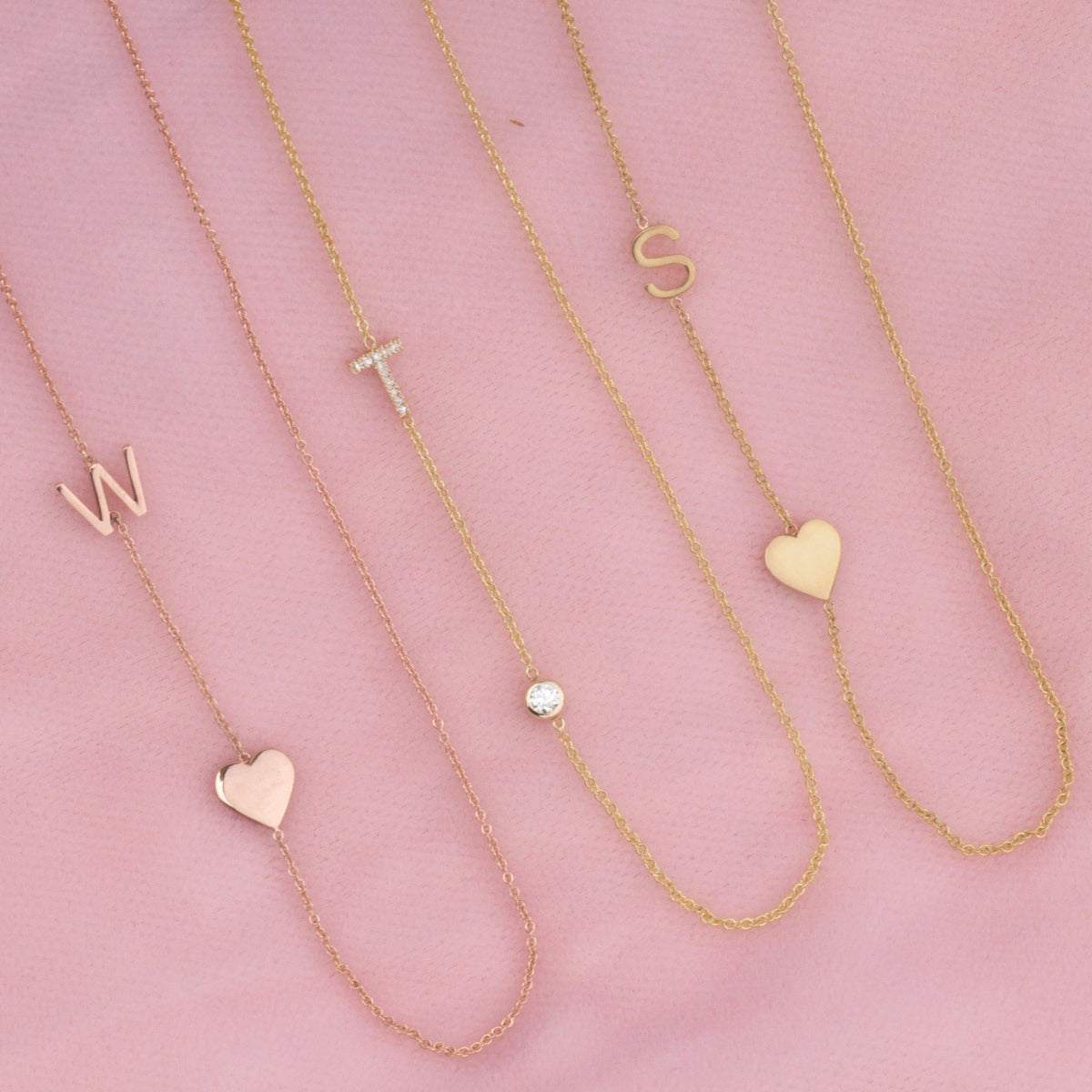 Three monogram necklaces against a pink background: a Monogram Necklace with Heart Rose Gold and a letter W charm, a Pavé Monogram Necklace with Diamond and a letter T charm in yellow gold and a Monogram Necklace with Heart Yellow Gold and a letter S charm.