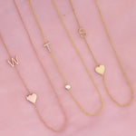Three monogram necklaces against a pink background: a Monogram Necklace with Heart Rose Gold and a letter W charm, a Pavé Monogram Necklace with Diamond and a letter T charm in yellow gold and a Monogram Necklace with Heart Yellow Gold and a letter S charm.