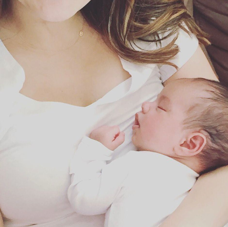 A woman holding a baby and wearing a gold necklace with a B letter charm. 
