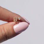 A Pavé Monogram Earring Set in the letter B being held by a hand with light pink nails against a light grey background. 