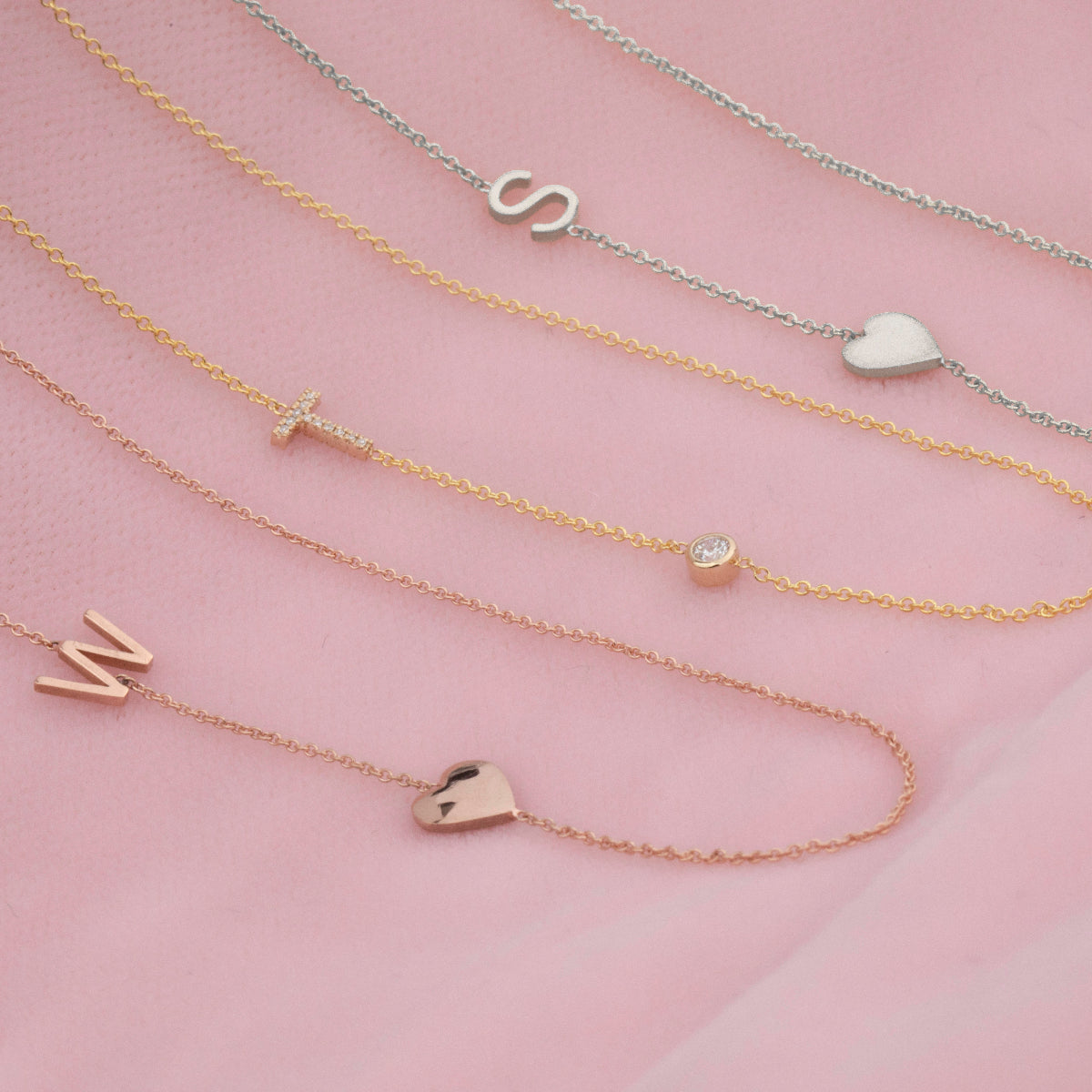Three necklaces laid on a pink fabric background: a Monogram Necklace with Heart Rose Gold and a letter W charm, a Pavé Monogram Necklace with Diamond and a letter T charm, and a Monogram Necklace with Heart White Gold and a letter S charm.