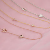 Three necklaces laid on a pink fabric background: a Monogram Necklace with Heart Rose Gold and a letter W charm, a Pavé Monogram Necklace with Diamond and a letter T charm, and a Monogram Necklace with Heart White Gold and a letter S charm.