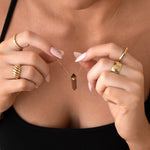 Close-up of a woman's décolletage wearing the Courage Retreat Necklace, with her hands holding the necklace and wearing an Etched Paris Ring, a Thick Band, a Geo Stacking Ring Square, and the Evermore Ring, styled in a black singlet top.