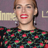 Busy Philipps wearing Star Studs, drop pearl earrings, and a black dress with red flowers against a dark purple background.