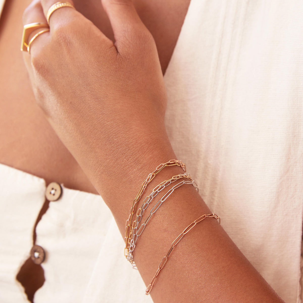 A close-up of a woman's wrist and hand wearing mixed metal Element Short Link Bracelets and Element Long Link Bracelets, and gold rings, styled with a white top. 