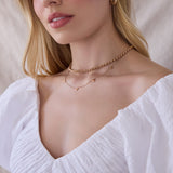 Close-up of a woman's décolletage wearing a Rolo Necklace and a 5 of Hearts Necklace in rose gold, with a white top and her blonde hair visible against a light neutral background.
