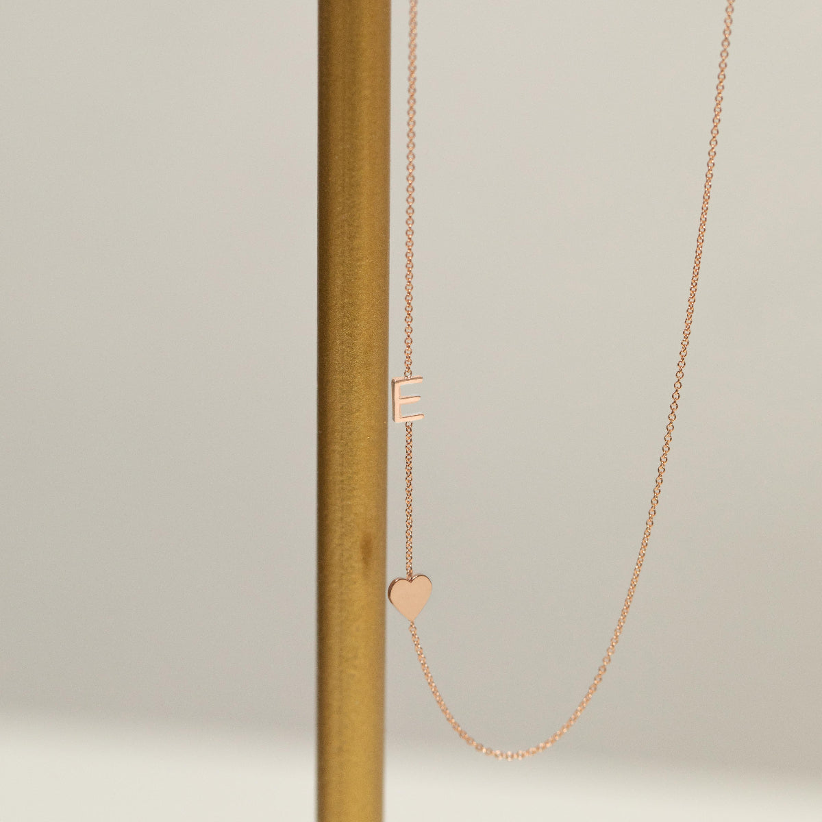 A Monogram Necklace with Heart Rose Gold and a letter E charm next to a brown stick against a light grey background. 