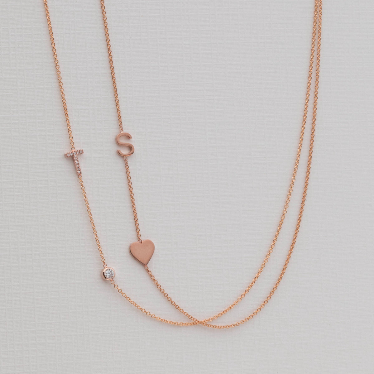 A Pavé Monogram Necklace with Diamond and a letter T charm in rose gold, and a Monogram Necklace with Heart Rose Gold and a letter S charm, against a neutral background.