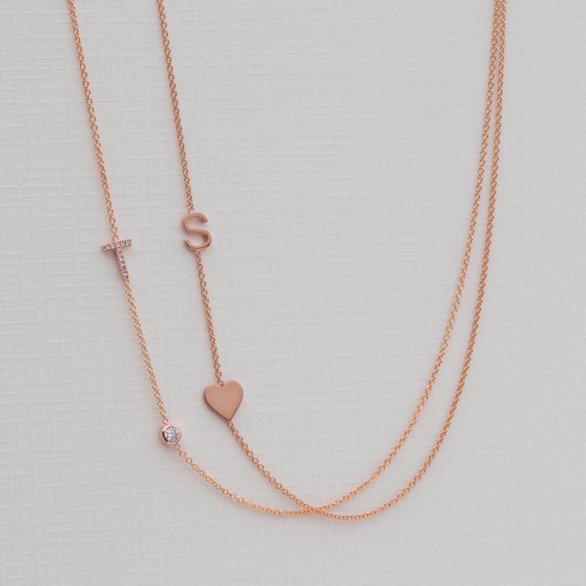 A Pavé Monogram Necklace with Diamond and a letter T charm in rose gold, and a Monogram Necklace with Heart Rose Gold and a letter S charm, against a neutral background.