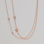 A Pavé Monogram Necklace with Diamond and a letter T charm in rose gold, and a Monogram Necklace with Heart Rose Gold and a letter S charm, against a neutral background.