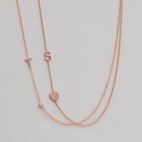 A Pavé Monogram Necklace with Diamond and a letter T charm in rose gold, and a Monogram Necklace with Heart Rose Gold and a letter S charm, against a neutral background.