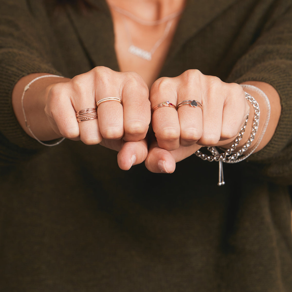 A pair of hands in fists wearing rose gold Delicate Trio Stacking Rings, two Pave Diamond Rings, two Birthstone Rings, a white gold Heartfelt Ring and Delicate Band, along with two white gold Rolo Bracelets and a Down the Line Bracelet, styled with a dark khaki green jumper. 