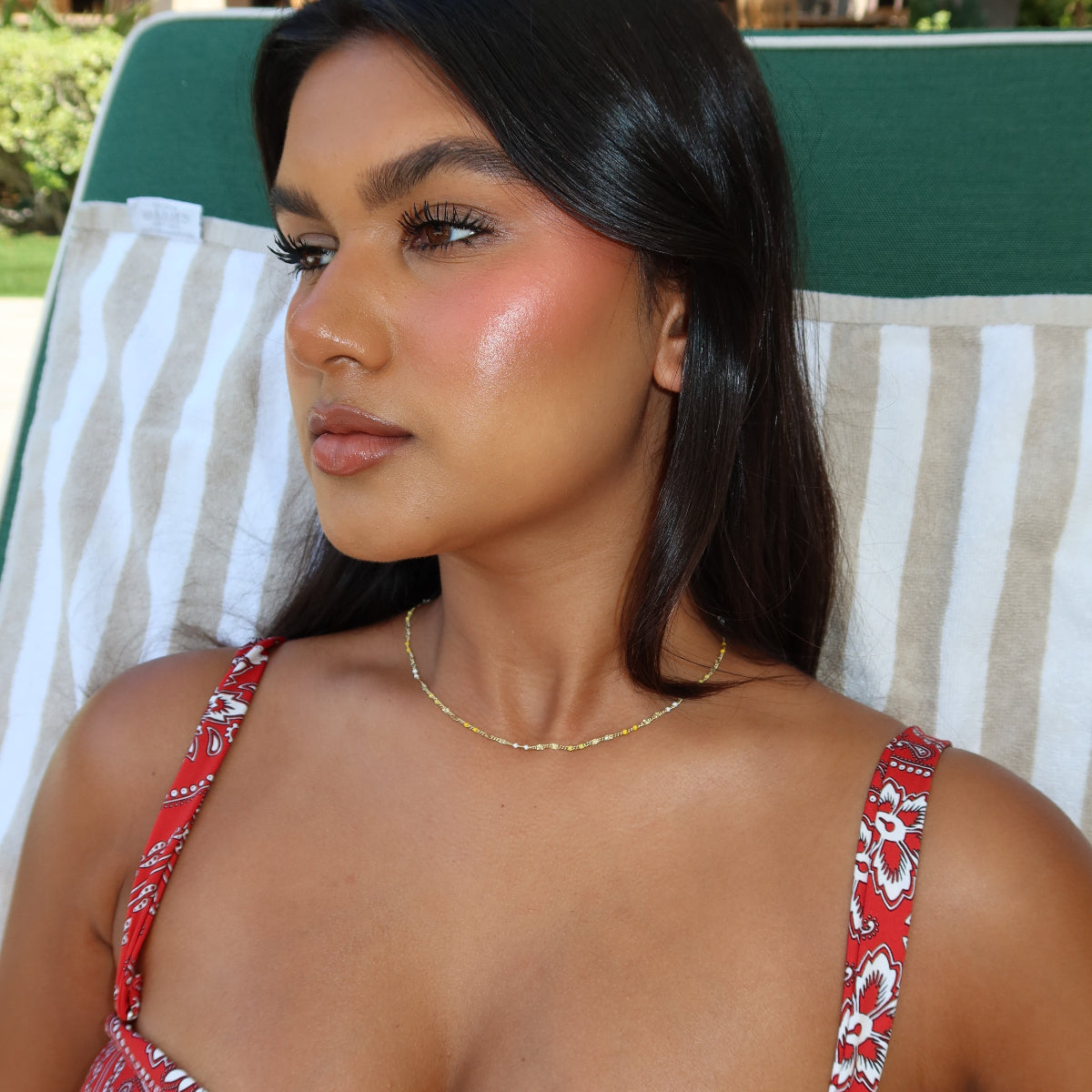 A woman wearing a Confetti Necklace in yellow, styled in a red floral bikini, laying on a green chair with a striped beige and white towel.
