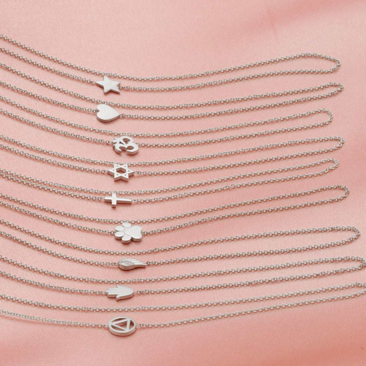 Nine white gold 14k Gold Asymmetrical Charm Necklaces with cable chains (star, heart, om, star of david, cross, paw, angel wing, hamsa and recovery) laid vertically on a light pink fabric background.