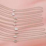 Nine white gold 14k Gold Asymmetrical Charm Necklaces with cable chains (star, heart, om, star of david, cross, paw, angel wing, hamsa and recovery) laid vertically on a light pink fabric background.