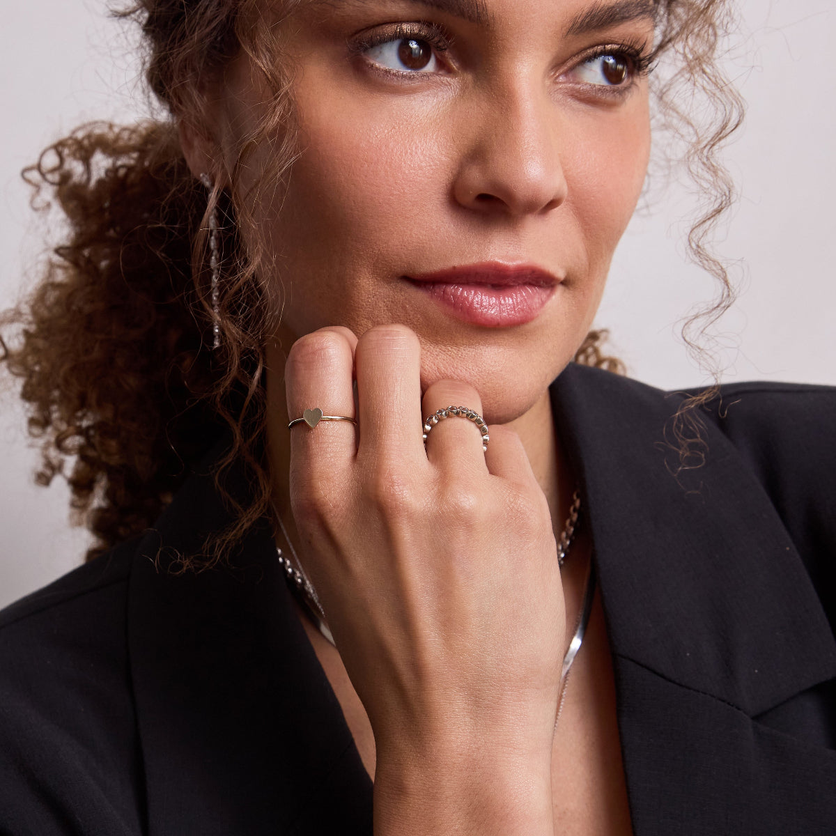 A woman wearing a white gold Heartfelt Ring, Bouquet Ring, Rolo Necklace and Sequence Chain, with brown curly hair and a black blazer against a light grey background. 